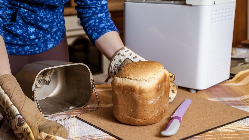 Eine Person hat gerade mit behandschuhten Händen ein frisch gebackenes Weißbrot aus dem Backbehälter eines Brotbackautomaten geholt. Das liegt auf einem Brett mit einem Messer neben dem Automaten.