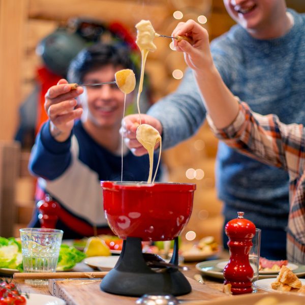 Auf einem Tisch steht ein Käsefondue-Set. Drumherum sind drei Personen, die auf ihren Gabeln Brot und flüssigen Käse balancieren.