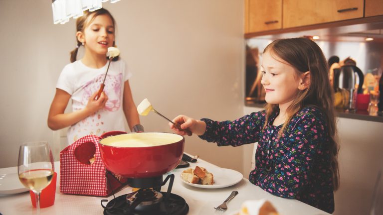 Zwei Kinder stehen um ein Käsefondue, in das sie Brotstücke tunken.