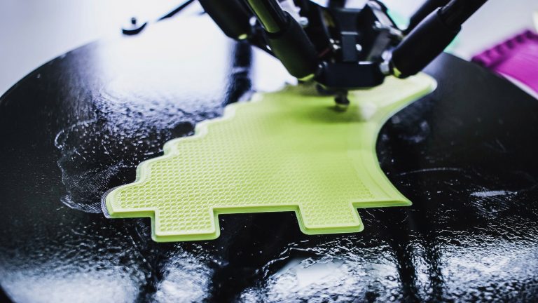 Makro-Ansicht einer 3D-Drucker-D&uuml;se, die gerade eine hellgr&uuml;ne Kunststoffschicht mit sichtbarer Wabenstruktur (Infill) auf ein schwarzes Druckbett auftr&auml;gt.