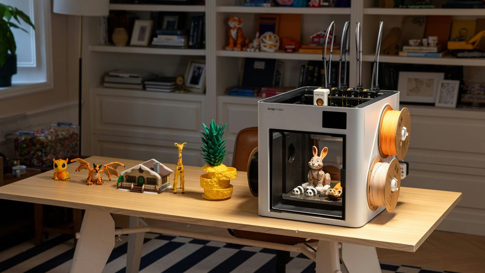 Ein wei&szlig;er 3D-Drucker steht auf einem Schreibtisch neben verschiedenen farbigen 3D-Druck-Objekten wie einer gelben Giraffe, einem kleinen Haus und Dinosaurier-Figuren.