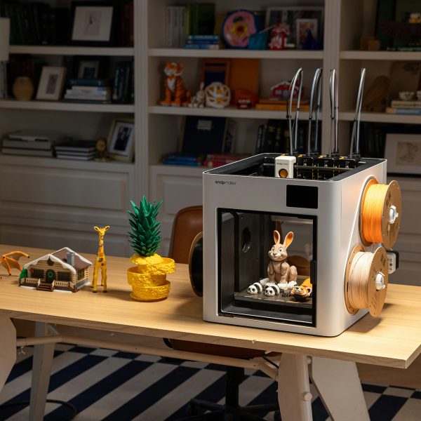 Ein wei&szlig;er 3D-Drucker steht auf einem Schreibtisch neben verschiedenen farbigen 3D-Druck-Objekten wie einer gelben Giraffe, einem kleinen Haus und Dinosaurier-Figuren.