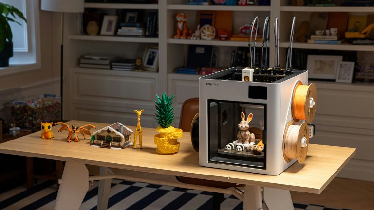 Ein wei&szlig;er 3D-Drucker steht auf einem Schreibtisch neben verschiedenen farbigen 3D-Druck-Objekten wie einer gelben Giraffe, einem kleinen Haus und Dinosaurier-Figuren.