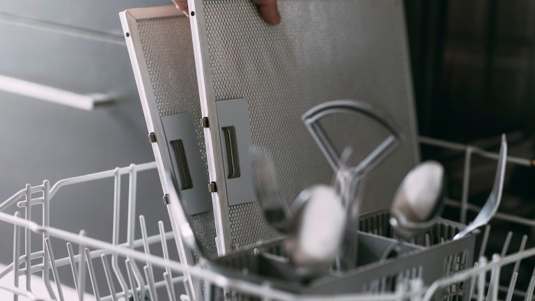 Dunstabzugshauben, Wie du das passende Modell für deine Küche findest Eine Person stellt zwei Metallfilter einer Abzugshaube in den unteren Korb einer geöffneten Spülmaschine.