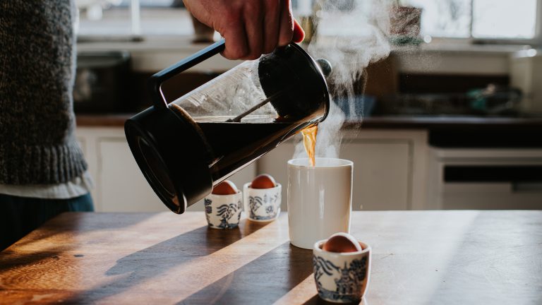 Die Hand einer Person sch&uuml;ttet dampfenden Kaffee aus einer French Press in eine Kaffeetasse, die auf der K&uuml;chen-Arbeitsplatte steht.
