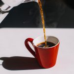 Eine rote Kaffeetasse steht auf einer wei&szlig;en Arbeitsplatte. Aus einer Mokkakanne wird Kaffee in die Tasse gegossen.
