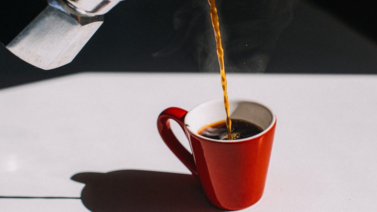 Eine rote Kaffeetasse steht auf einer wei&szlig;en Arbeitsplatte. Aus einer Mokkakanne wird Kaffee in die Tasse gegossen.