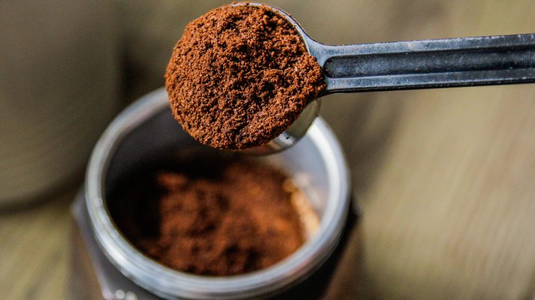 Fokus auf einen Kaffeel&ouml;ffel, der geh&auml;uft ist mit Kaffee, um diesen in das Sieb einer Mokkakanne zu geben. Diese ist leicht verschwommen im Hintergrund zu sehen.