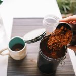Eine Person sch&uuml;ttet frisch gemahlenen Kaffee in eine Kaffeedose, die auf einer Anrichte neben einer Kaffeetasse steht.&nbsp;