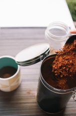 So lagerst du Kaffeepulver und Bohnen richtig, 11 Tipps für volles Aroma Eine Person schüttet frisch gemahlenen Kaffee in eine Kaffeedose, die auf einer Anrichte neben einer Kaffeetasse steht.