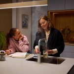 Zwei Personen in moderner K&uuml;che. Eine Person gie&szlig;t Wasser aus einem Quooker-Wasserhahn in ein Glas.