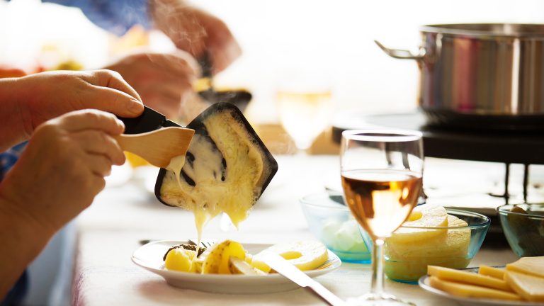 Eine Person schabt an einem Esstisch mit einem Holzspatel K&auml;se aus einem Raclettepf&auml;nnchen auf einen Teller.