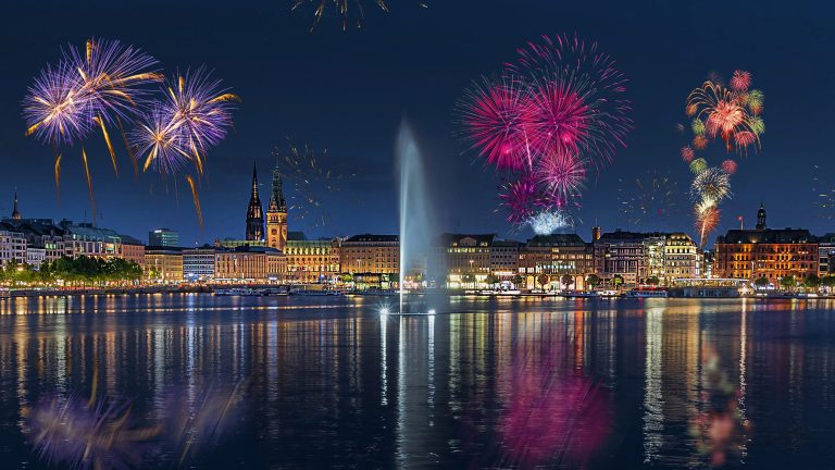 Eine Weitwinkelaufnahme mit Blick auf die Wasserfont&auml;ne und die Hamburgerbinnenalster in Richtung Jungfernstieg mit einem Feuerwerk dar&uuml;ber.