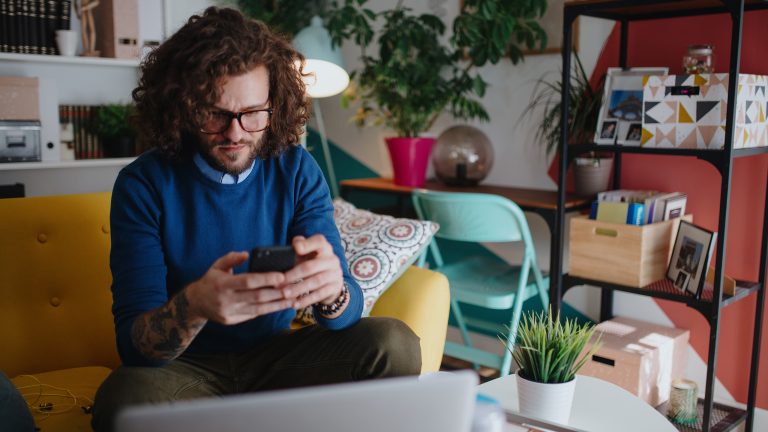 Eine Person mit lockigem Haar sitzt auf einer Couch und blickt konzentriert auf ihr Smartphone, w&auml;hrend ein Laptop daneben steht.