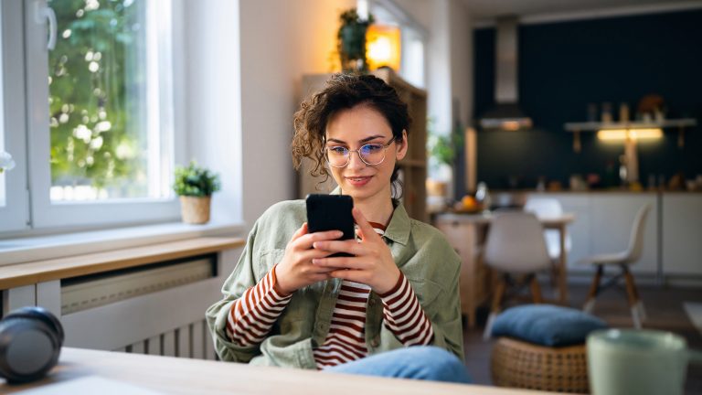 Eine l&auml;chelnde Person sitzt zu Hause an einem Tisch und h&auml;lt ihr Smartphone.