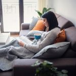 Eine Person sitzt auf einer Couch und liest in einem Buch. Auf ihrem Scho&szlig; liegt eine Heizdecke.