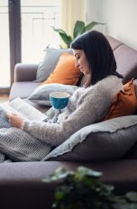 Eine Person sitzt auf einer Couch und liest in einem Buch. Auf ihrem Scho&szlig; liegt eine Heizdecke.