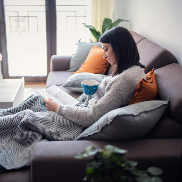 Eine Person sitzt auf einer Couch und liest in einem Buch. Auf ihrem Scho&szlig; liegt eine Heizdecke.