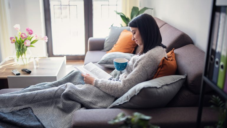 Eine Person sitzt auf einer Couch und liest in einem Buch. Auf ihrem Scho&szlig; liegt eine Heizdecke.