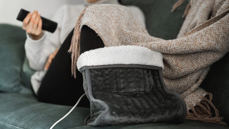 Heizdecken und Heizkissen, So hast du es im Winter wohlig warm Im Fokus des Fotos ist ein elektrischer Fußwärmer zu sehen, der die Füße einer, auf der Couch sitzenden Person, wärmt.