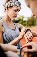 Drei Menschen stehen an einer Laufbahn. Im Fokus steht eine Person, die auf eine Smartwatch an ihrem linken Handgelenk schaut.