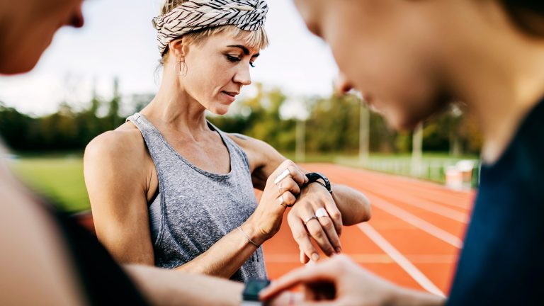Drei Menschen stehen an einer Laufbahn. Im Fokus steht eine Person, die auf eine Smartwatch an ihrem linken Handgelenk schaut.