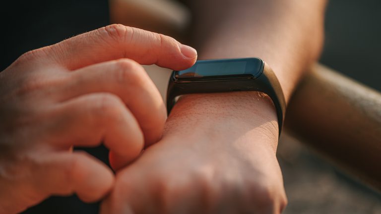 Detailaufnahme eines linken Handgelenks an dem eine Fitnesstracker getragen wird. Der Zeigefinger der rechten Hand bedient diesen.