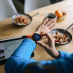 Vogelperspektive auf einen Fr&uuml;hst&uuml;ckstisch mit M&uuml;slisch&uuml;sseln und Kaffeetassen. Im Fokus des Bildes ist der Arm einer Person, die am Tisch sitzt und eine Smartwatch am linken Handgelenk tr&auml;gt.