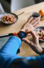 Fitness-Tracker oder Smartwatch, Was sind die Unterschiede? Vogelperspektive auf einen Frühstückstisch mit Müslischüsseln und Kaffeetassen. Im Fokus des Bildes ist der Arm einer Person, die am Tisch sitzt und eine Smartwatch am linken Handgelenk trägt.
