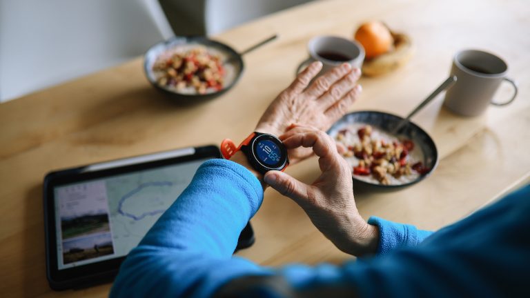Vogelperspektive auf einen Fr&uuml;hst&uuml;ckstisch mit M&uuml;slisch&uuml;sseln und Kaffeetassen. Im Fokus des Bildes ist der Arm einer Person, die am Tisch sitzt und eine Smartwatch am linken Handgelenk tr&auml;gt.