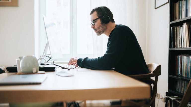 Eine Person sitzt im Homeoffice an einem gro&szlig;en Holzschreibtisch vor einem Computer. Dabei tr&auml;gt sie Kopfh&ouml;rer und tippt.