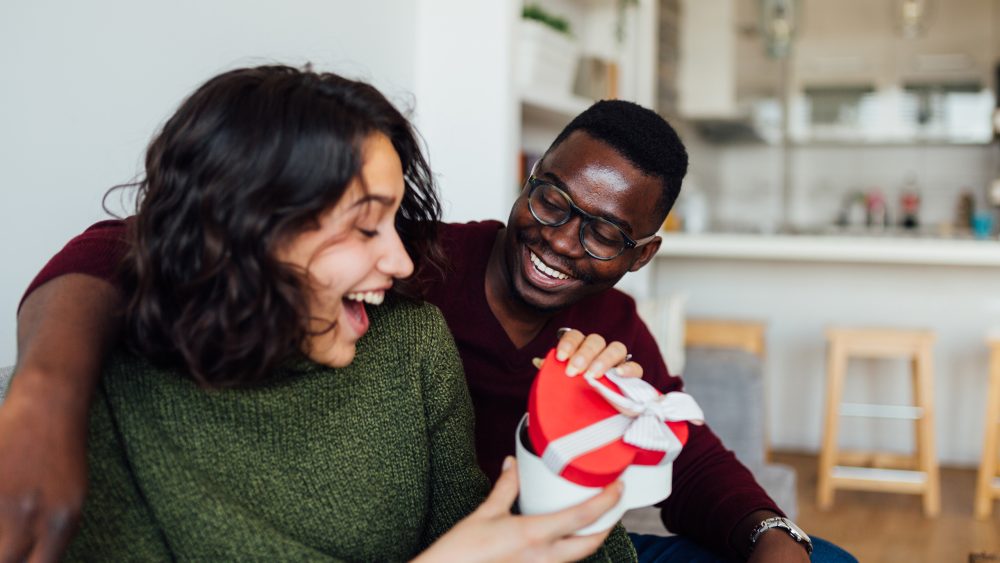 Zwei Personen sitzen auf einer Couch in einer offenen Wohnk&uuml;che. Eine Person hat den Arm um die andere gelegt, w&auml;hrend diese ein herzf&ouml;rmiges Geschenk &ouml;ffnet und sich freut.