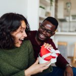 Zwei Personen sitzen auf einer Couch in einer offenen Wohnk&uuml;che. Eine Person hat den Arm um die andere gelegt, w&auml;hrend diese ein herzf&ouml;rmiges Geschenk &ouml;ffnet und sich freut.