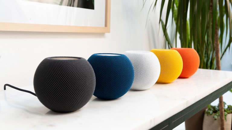 F&uuml;nf Apple HomePod mini in unterschiedlichen Farben stehen in einer Reihe auf einer wei&szlig;en Marmoranrichte an einer Wand. Im Hintergrund ist der Anschnitt von einem gerahmten Bild und eine Palme.