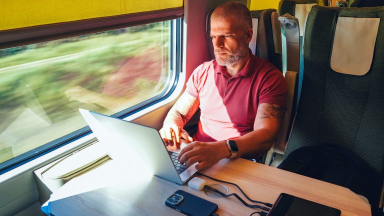 Eine Person sitzt in einem Vierer in einem Zug und hat einen Laptop vor sich, neben dem eine gro&szlig;e Powerbank liegt, an die auch noch ein Handy und ein Headphone-Case angeschlossen sind.