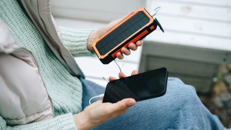 Eine Person sitzt im Freien und h&auml;lt ein Smartphone und eine Solar-Powerbank in beiden H&auml;nden. Sie sind mit einem Kabel verbunden.