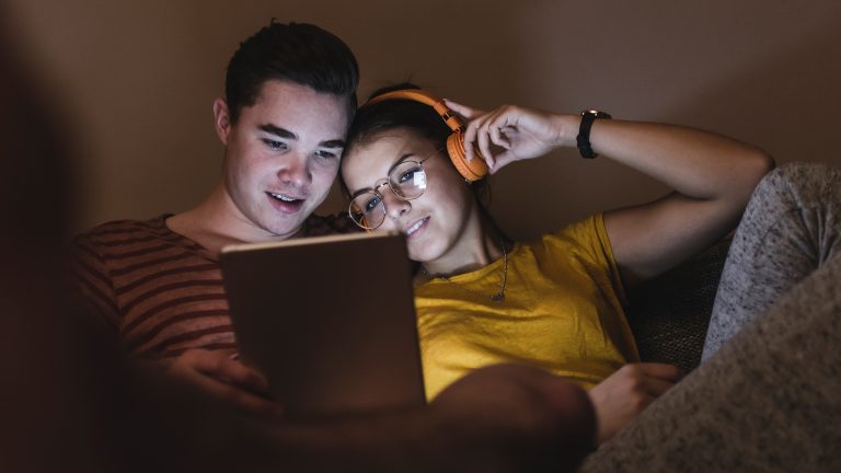 Zwei Personen sitzen aneinandergelehnt auf einer Couch und schauen auf ein Tablet, von dem sie angeleuchtet werden. Eine Person tr&auml;gt Over-Ear-Kopfh&ouml;rer.