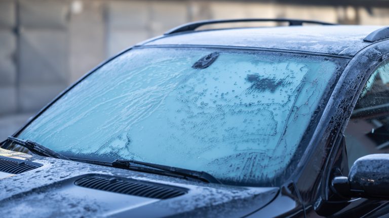 Winter‑Gadgets für eine angenehm-kalte Jahreszeit Ausschnitt auf die Motorhaube und die Frontscheibe eines schwarzen Autos, welches mit einer Eisschicht überzogen ist.