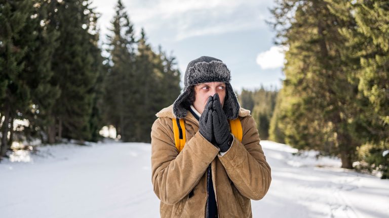 Eine Person steht in einer winterlichen Landschaft. Sie tr&auml;gt warme Kleidung, eine M&uuml;tze und Handschuhe an den H&auml;nden, die sie sich vor den Mund h&auml;lt, um sie warm anzublasen.