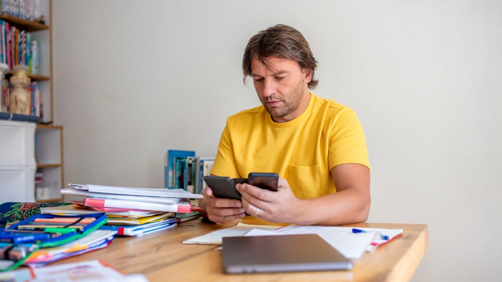 Eine Person sitzt mit zwei Smartphones in der Hand an einem Tisch mit Stiften und einem Laptop.