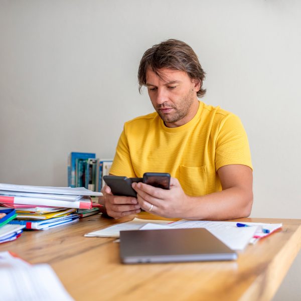 Eine Person sitzt mit zwei Smartphones in der Hand an einem Tisch mit Stiften und einem Laptop.