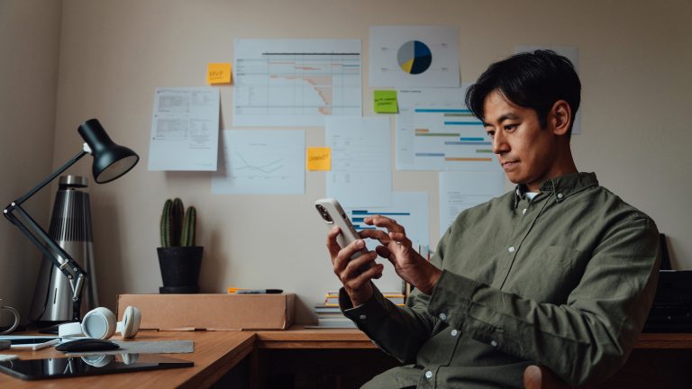 Authenticator auf neues Handy übertragen, So geht es Eine Person sitzt in einem Büro und hält ein Smartphone in der Hand. Im Hintergrund sind Graphen an einer Wand zu sehen.