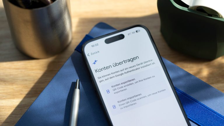 Authenticator auf neues Handy übertragen, So geht es Ein iPhone liegt auf einem Notizbuch. Auf dem Display ist die Konto-Übertragung des Google Authenticators zu sehen.