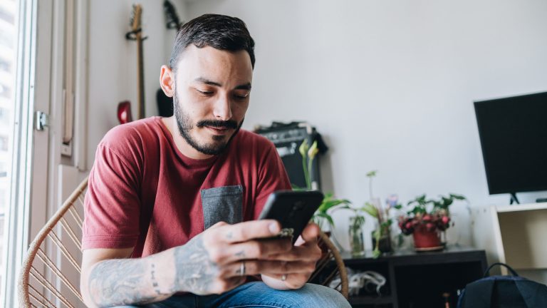Eine Person sitzt auf einem Korbstuhl in einem Zimmer. Sie richtet sich nach vorn, hat ihre Ellenbogen auf den Knien und blickt auf ein Smartphone in ihren H&auml;nden.
