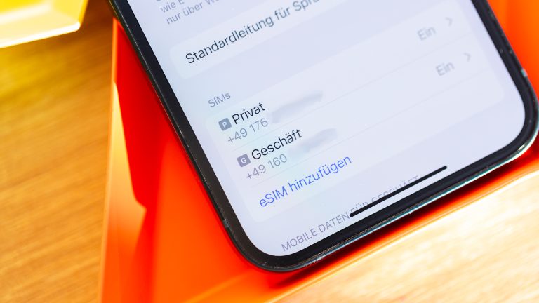 Der untere Teil eines Handys, das in einer eckigen Schale auf einem Holztisch liegt. Darauf zu sehen sind die Einstellungen auf einem Handy, wo eine Privat- und eine Gesch&auml;fts-Nummer zu sehen sind.