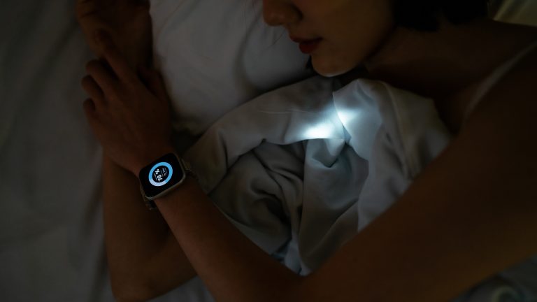 Eine Person liegt in der Seitenschl&auml;ferposition in einem halbdunklen Raum im Bett. Ein kleines Licht, das von einem Wearable kommt, scheint auf die Bettdecke.