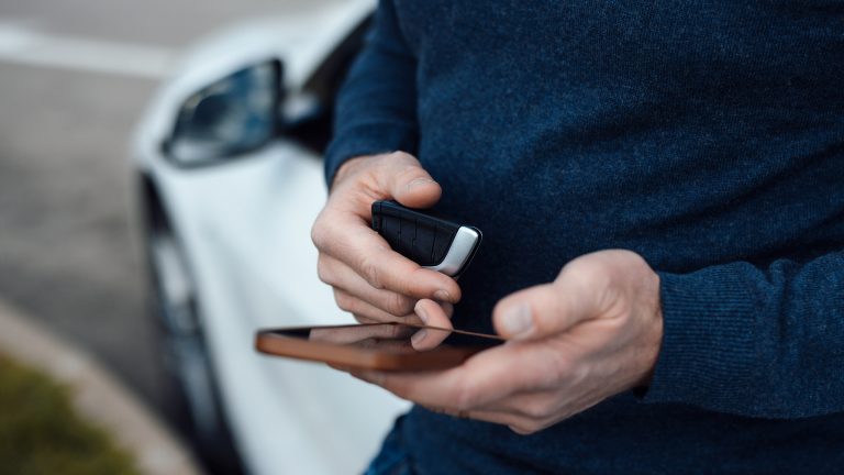 Digitaler Fahrzeugschein, Infos und Einrichtung Eine Person lehnt an einem Auto, hält den Fahrzeugschlüssel in der Hand und bedient mit der anderen ihr Smartphone.