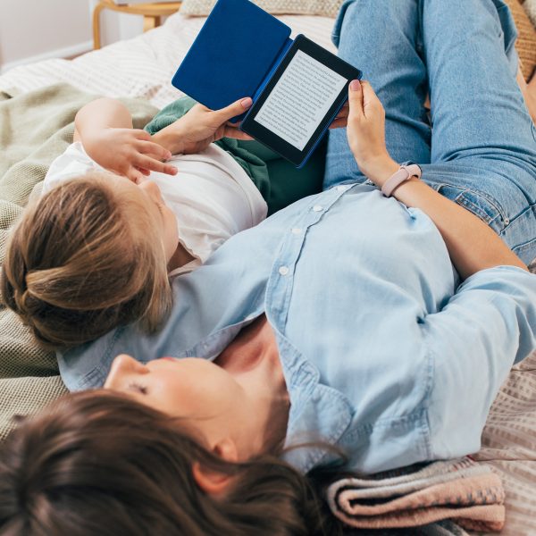 Eine Person liegt r&uuml;cklings auf einem Bett und h&auml;lt ein Kind im Arm, w&auml;hrend sie auf einem eReader ein Buch liest.