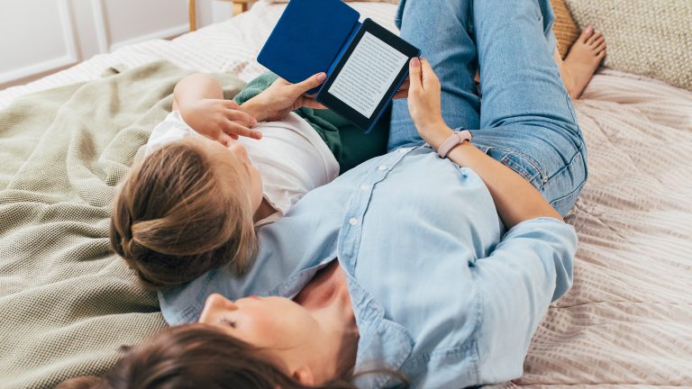 Eine Person liegt r&uuml;cklings auf einem Bett und h&auml;lt ein Kind im Arm, w&auml;hrend sie auf einem eReader ein Buch liest.