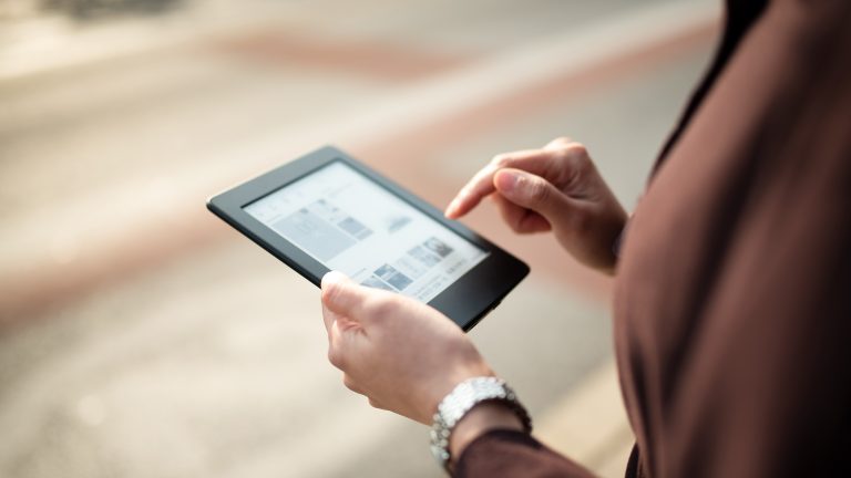 Tolino oder Kindle - Welche Lesewelt passt besser zu deinem Alltag? Eine Person steht im Freien und hält in der einen Hand einen eReader und tippt mit der anderen Hand darauf.